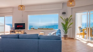 Ein modernes Wohnzimmer mit einer großen Couch, einem Fernseher, der 'NETFLIX' zeigt, einem großen Fenster mit Meerblick und einer Zimmerpflanze.