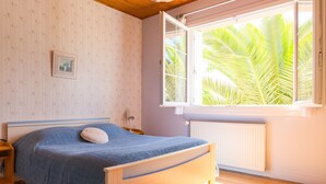 A bedroom with a bed, a window with a view of palm trees, a radiator, and a picture on the wall. - House for 6 - all on foot (Le Guilvinec)
