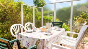 Un invernadero con una mesa dispuesta para comer, rodeado de sillas blancas y vistas a un jardín exuberante.