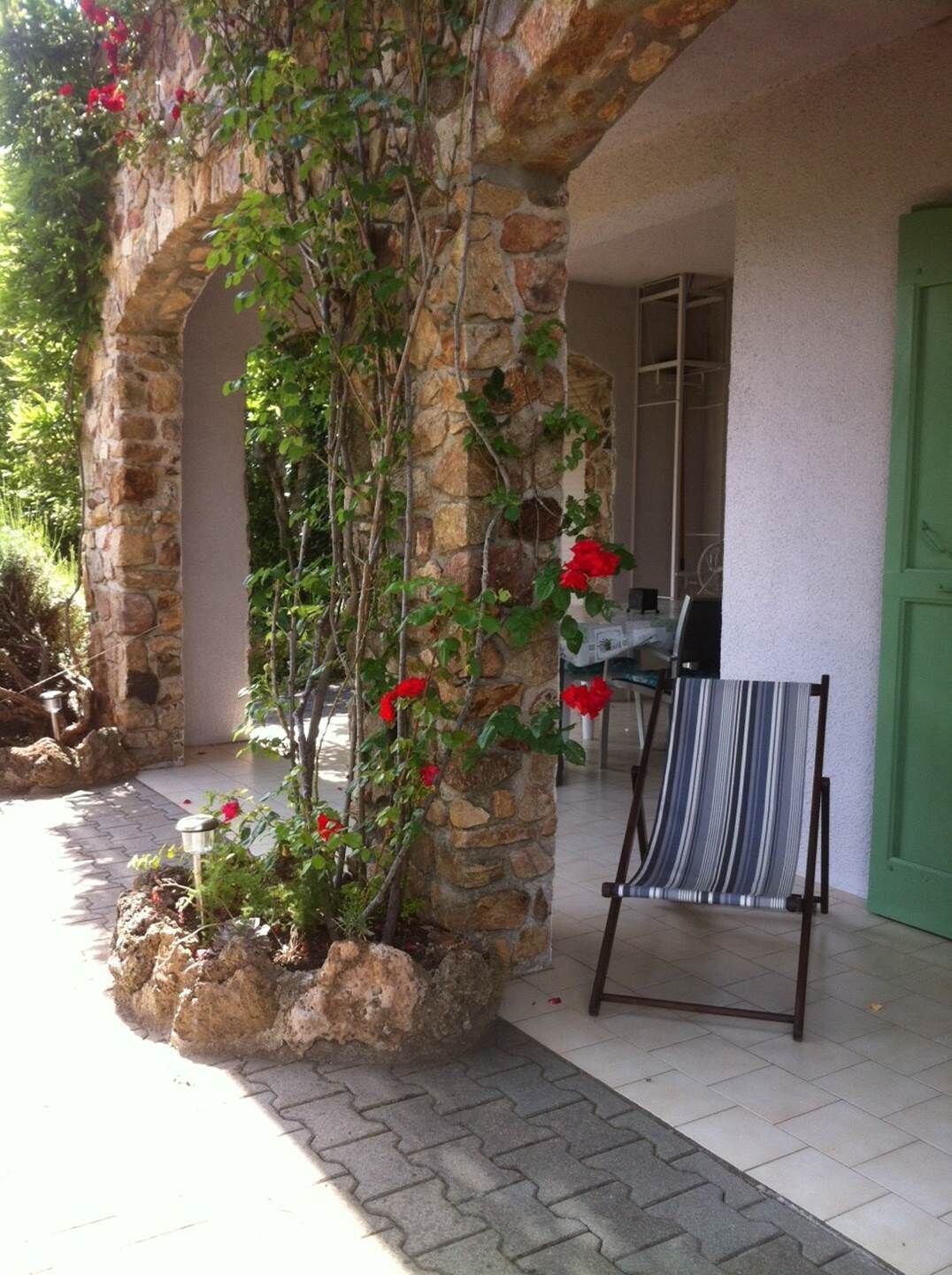 Un pilar de piedra con plantas trepadoras y flores rojas, una silla de playa a rayas y una puerta verde.