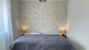Een tweepersoonsbed met een grijze deken, twee bedlampjes en behangpapier met een bladmotief.