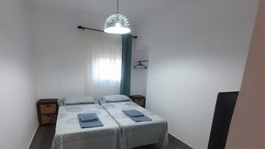 Une petite chambre propre, avec deux lits, une table de chevet, un luminaire suspendu et une fenêtre avec des rideaux.