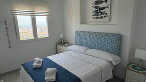 Habitación de hotel con cama, mesita de noche, lámpara y ventana con vista a edificios.