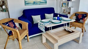 Una sala de estar con un sofá azul, sillas de mimbre, una mesa de centro de madera y una mesa de comedor con platos y vasos.