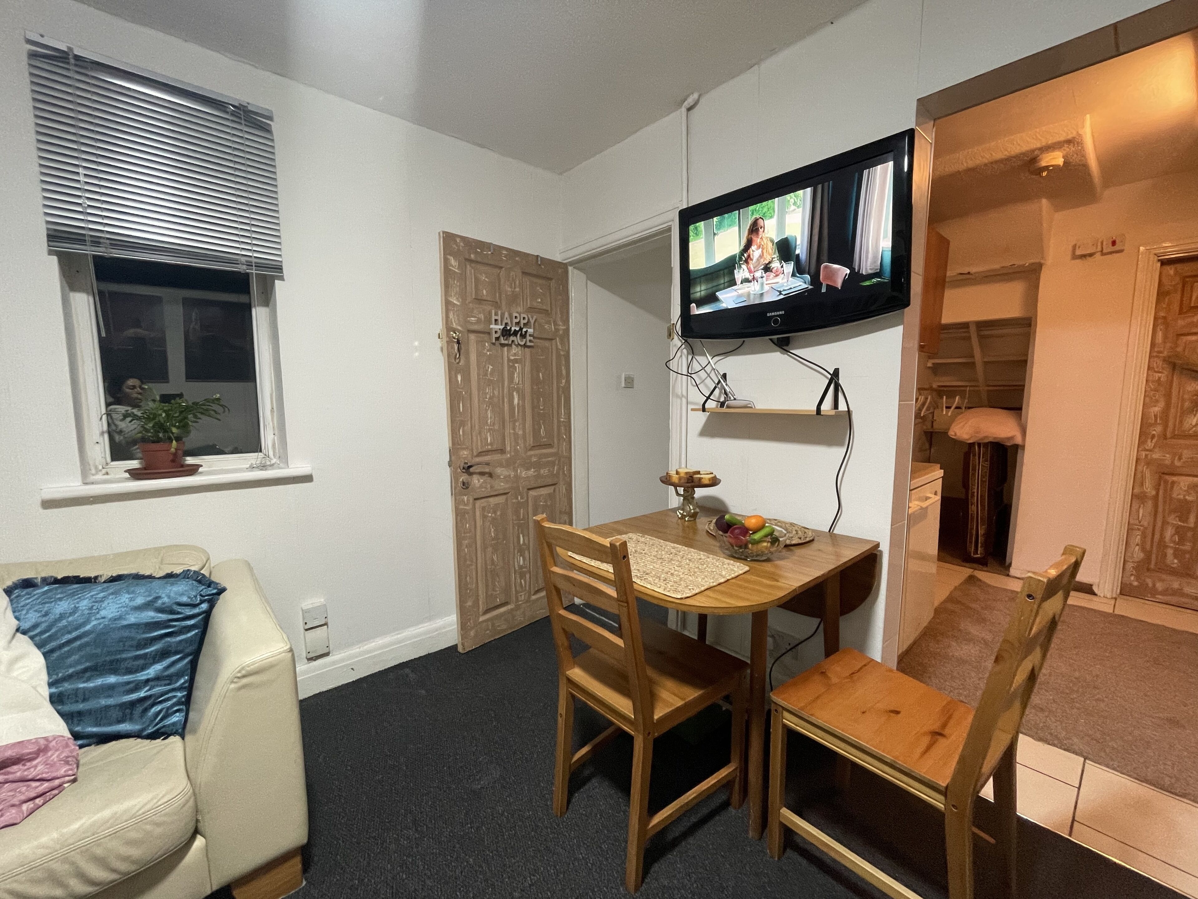 Um quarto pequeno e acolhedor com uma mesa de jantar de madeira, duas cadeiras, uma televisão na parede, uma janela com persianas e uma porta com o letreiro 'HAPPY PEACE'.