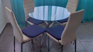 Mesa redonda de vidrio con cuatro sillas, cada una con un cojín azul, en una habitación con ventanas grandes y vistas a edificios.