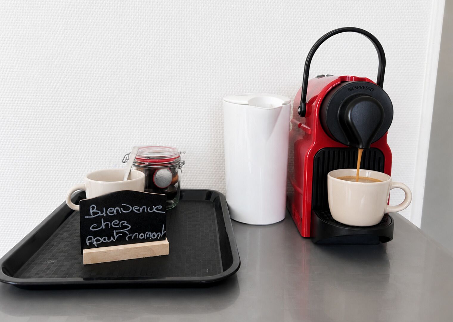 Una cafetera expreso roja sobre una bandeja, con una taza blanca y un frasco pequeño de café. Un letrero negro con texto blanco dice 'Bienvenue chez Apap Nomark'.