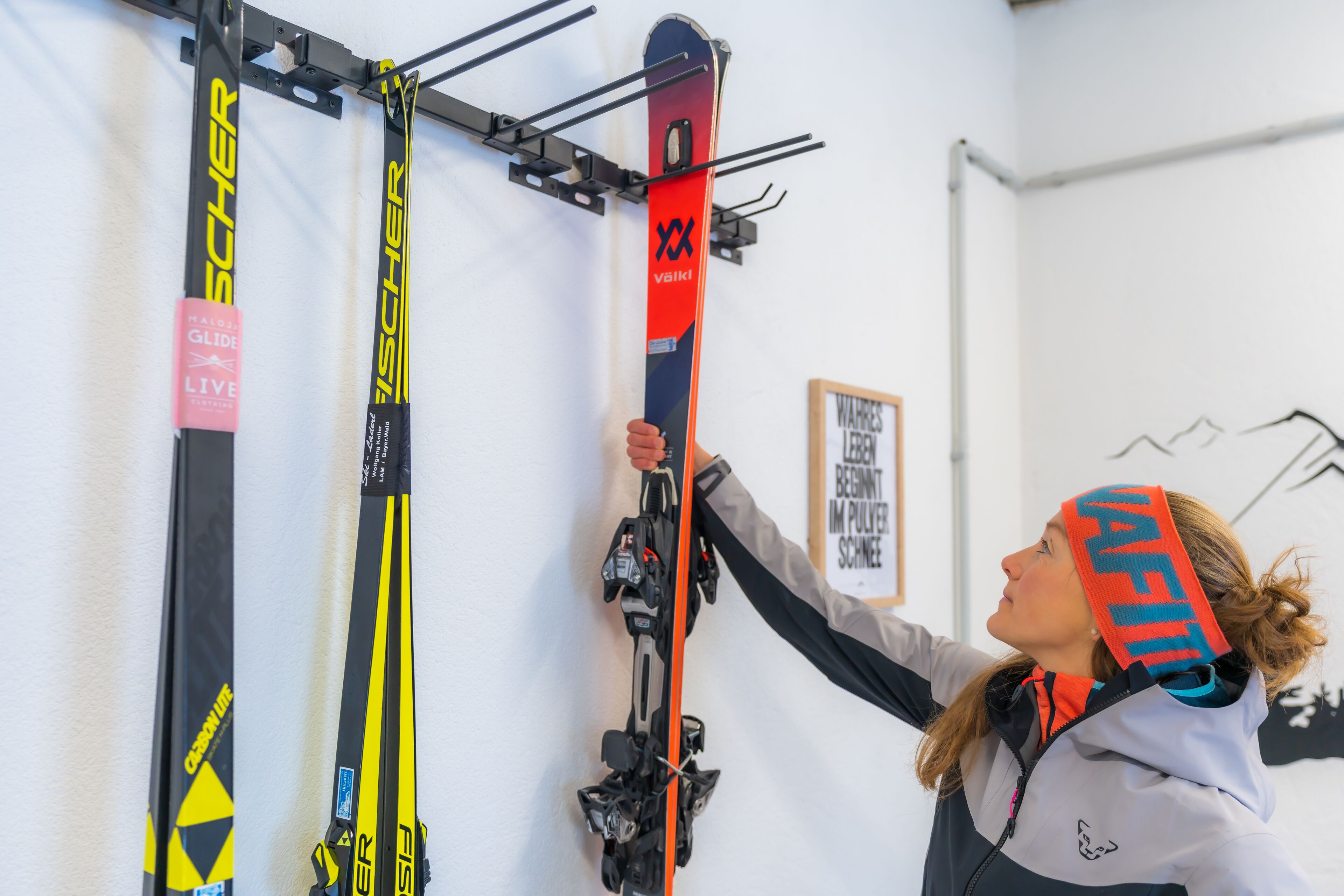 Eine Person hält ein Paar Ski, im Hintergrund ist ein Wandbefestigungssystem für Skier zu sehen.
