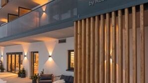 A modern building with a prominent 'Zeitlang by Rosenzwirt' sign. It features a wooden facade, outdoor seating area with a sofa and coffee table, and a snow-covered ground. - zeitlang by Rösslwirt (Lam)