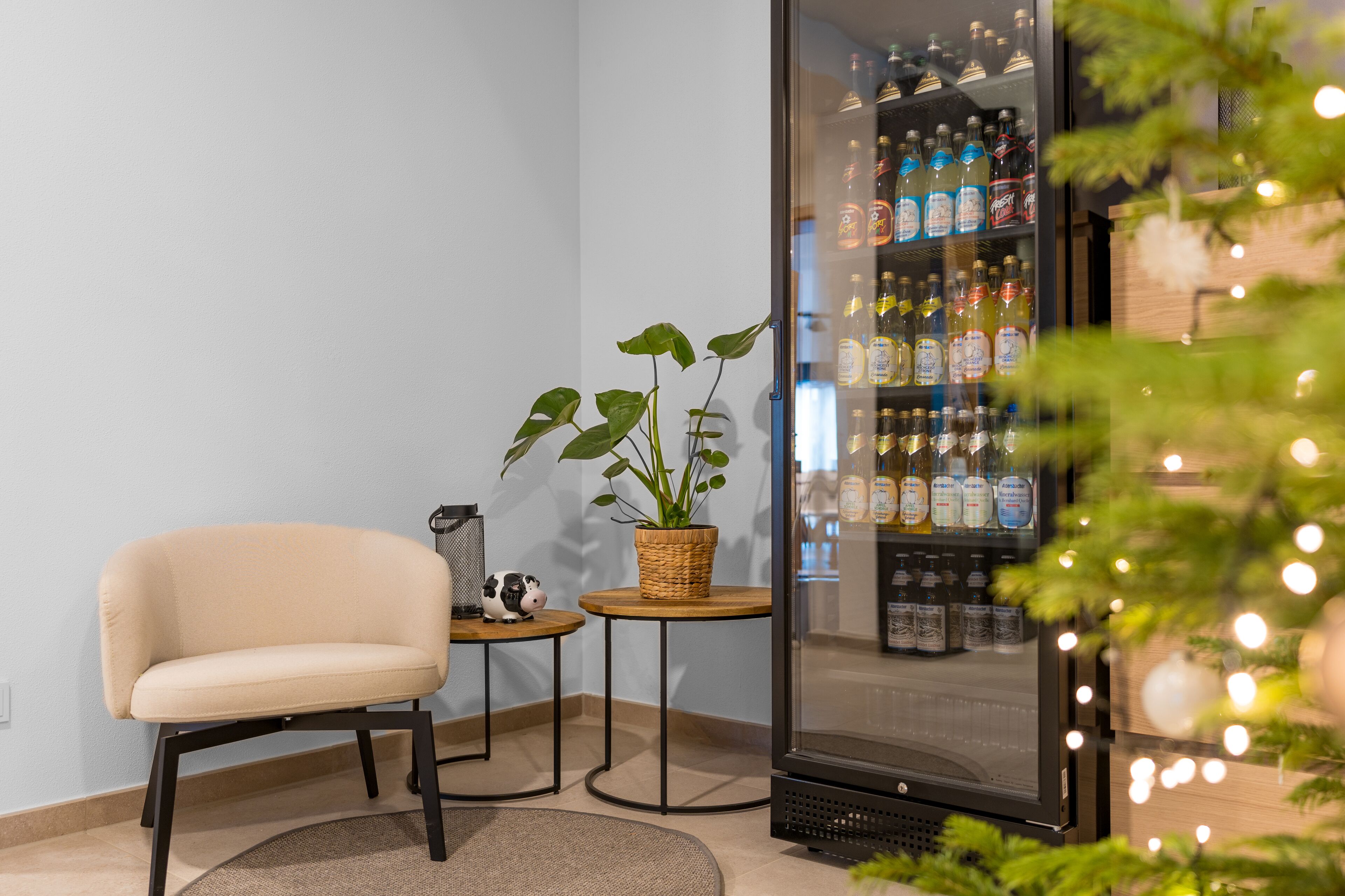 A corner of a room with a beige chair, a small round table with a plant, and a glass cabinet filled with bottles.