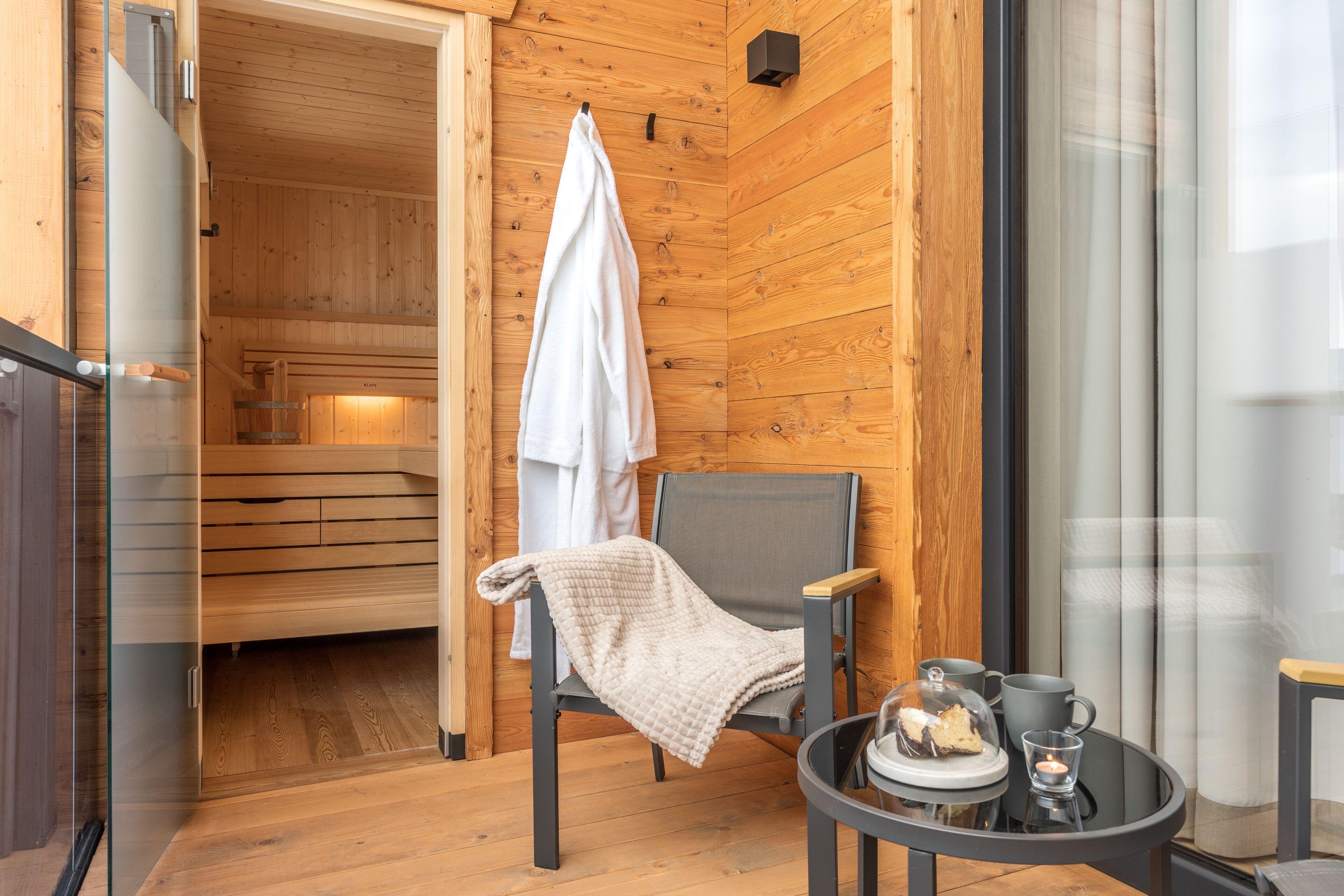 A wooden interior with a sauna, a glass door, a chair with a blanket, and a table with a cup and a plate.