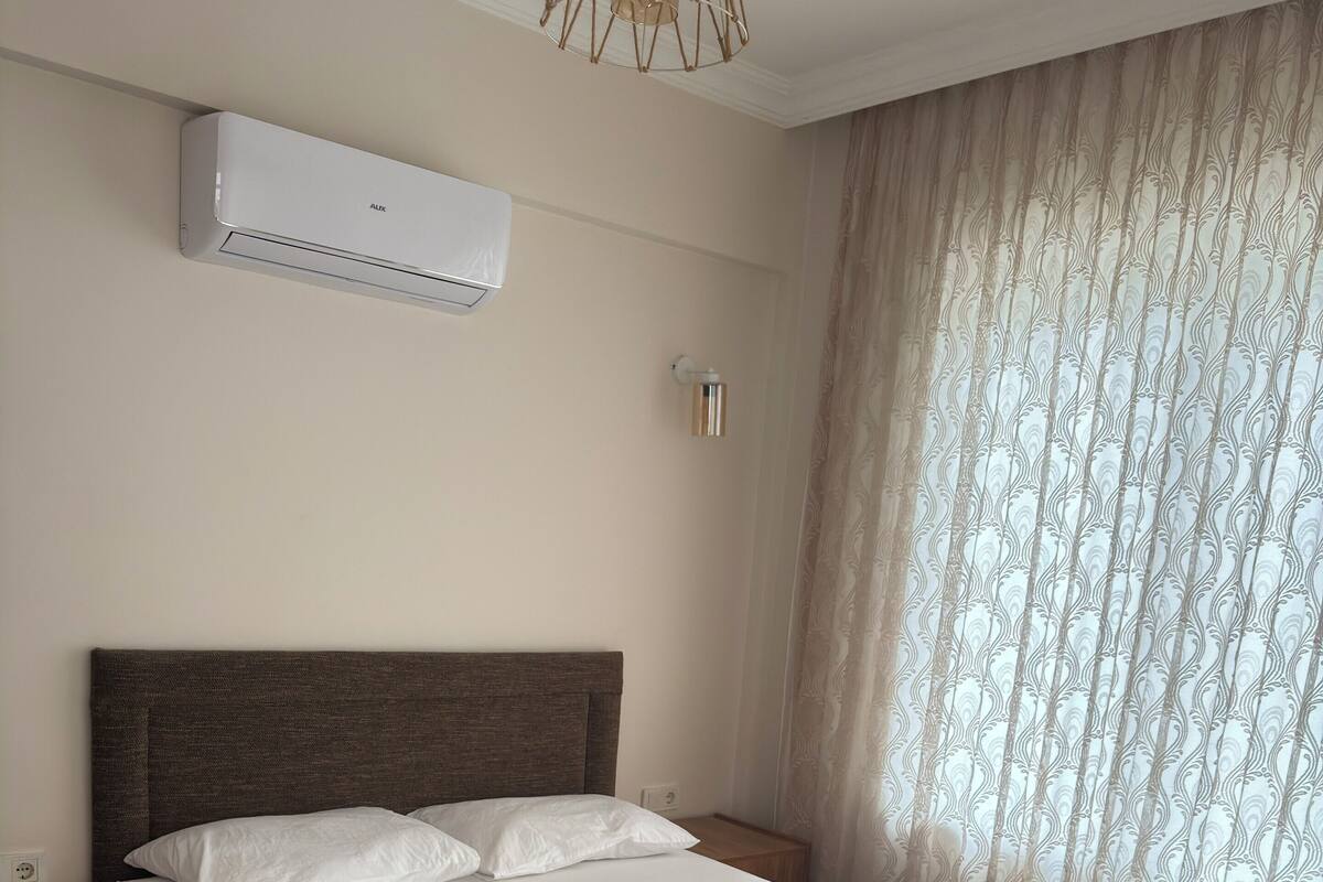 A neatly made bed with white linens and towels, a wooden headboard, a nightstand with a lamp, and a wall-mounted air conditioner.