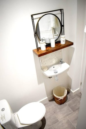 A bathroom with a white toilet, a wooden vanity with a sink, a mirror, and a small trash bin.