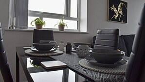 A modern dining area with a glass table, black chairs, and a black and white patterned table runner. There are potted plants on the windowsill and a framed artwork on the wall. - Remarkable 2-bed Apartment in Leafy Sefton Park (Liverpool)