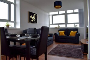 A modern living room with a dining area, featuring a dark blue sofa with yellow cushions, a dining table with chairs, and a large window with multiple panes.