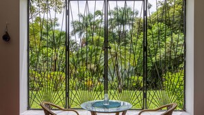Une pièce dotée d’une grande fenêtre donnant sur un jardin luxuriant. On y trouve une table ronde en verre, ornée d’un vase central, et deux chaises en osier.