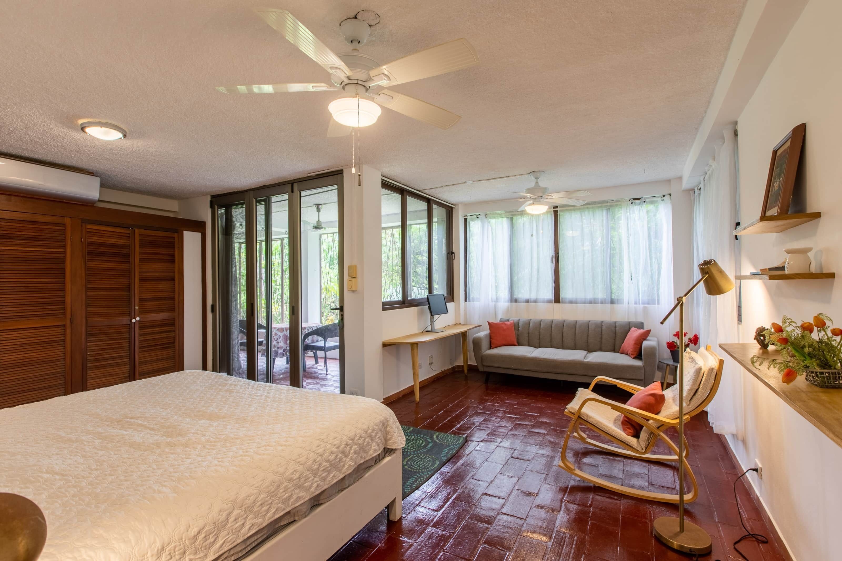 Un dormitorio con cama, una mecedora, un escritorio y una ventana con vistas a la vegetación.