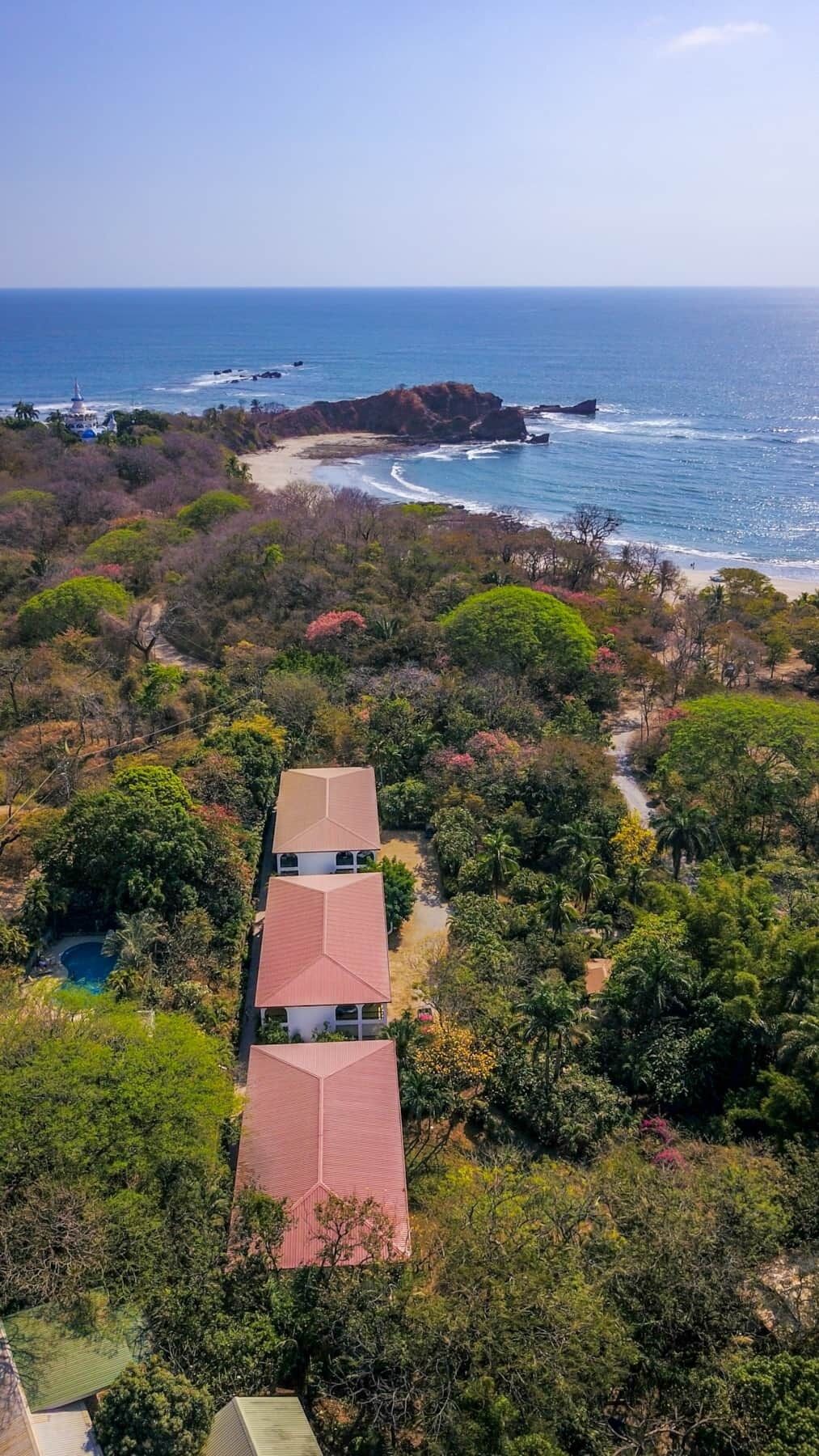 A two-story building with a red roof, surrounded by dense greenery and a beach with rocky outcrops.