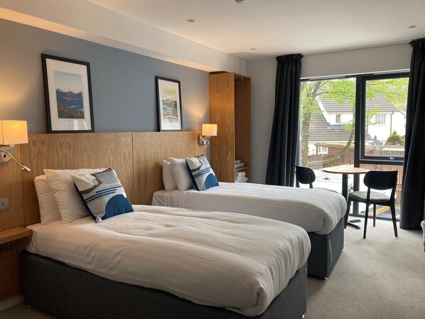 A hotel room with two beds, a small table, a chair, and a window with curtains.