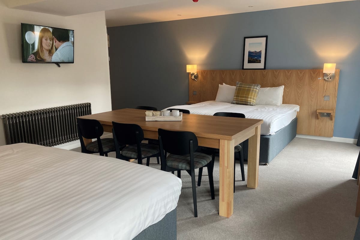 Una habitación de hotel con una cama grande, una cama más pequeña, una mesa de comedor con sillas, un radiador y un televisor montado en la pared.