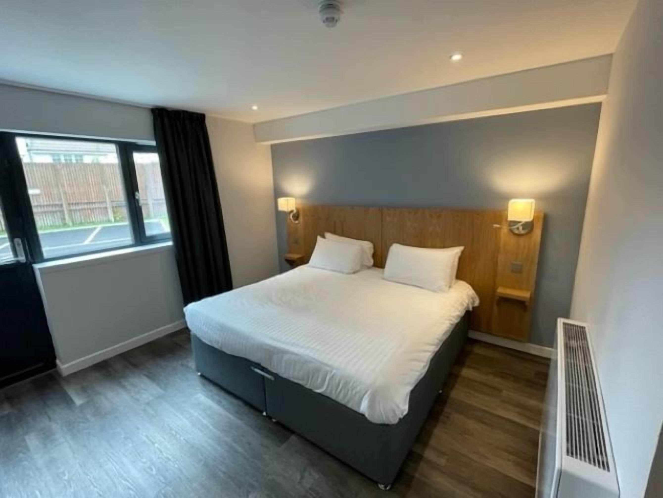 A modern hotel room with a large bed, two bedside tables with lamps, a window with curtains, and a radiator.