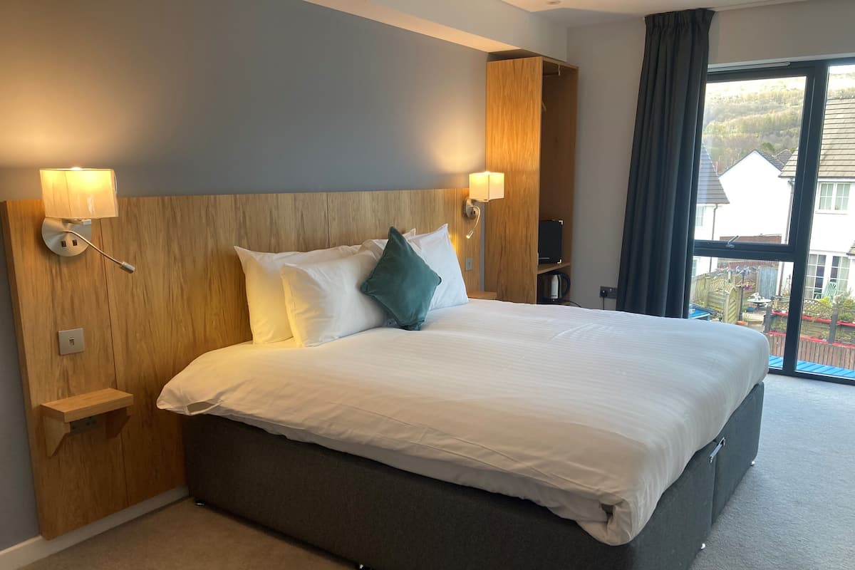 Habitación de hotel con una cama grande, cabecera de madera, dos lámparas de noche y vistas al exterior.