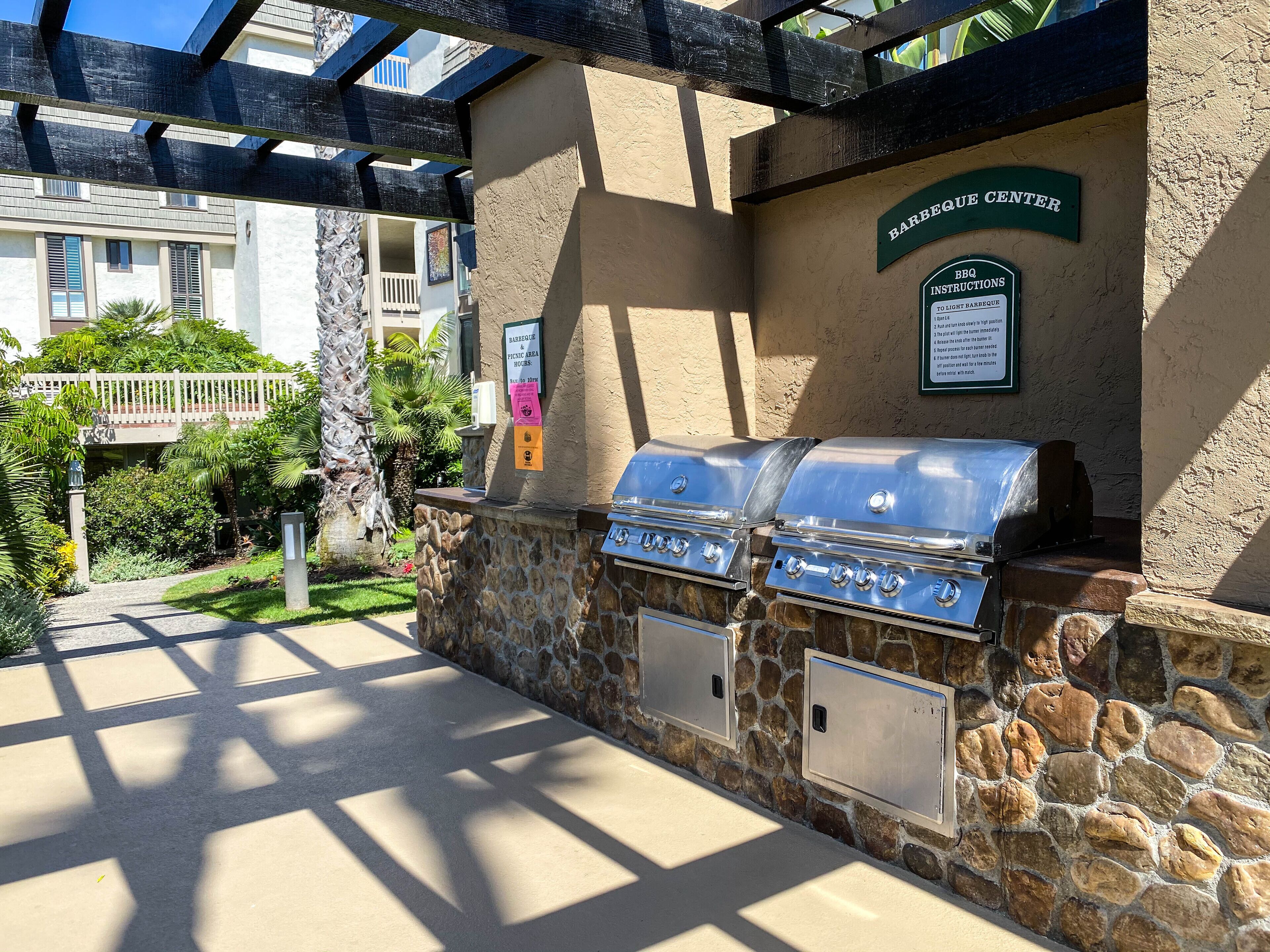 An outdoor barbecue area with stone walls and grilling stations, a sign indicating BBQ instructions, and a view of a building in the background.