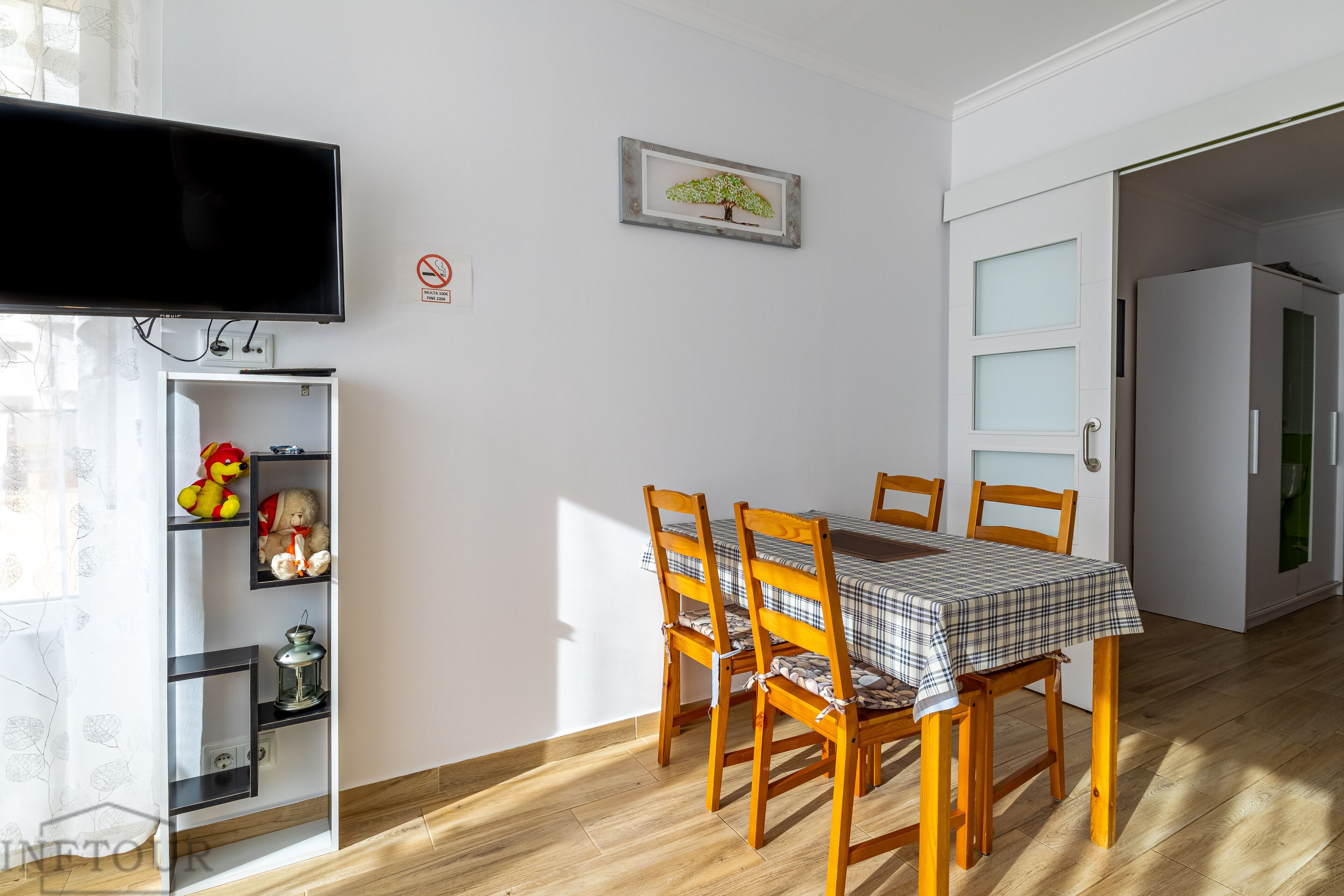 Un comedor con una mesa dispuesta para cuatro personas, sillas de madera, un televisor montado en la pared, un estante con juguetes de peluche y un cuadro enmarcado.