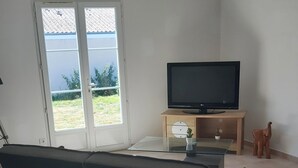 A living room with a sofa, a television on a wooden stand, a small table with a cactus, and a sliding glass door leading to a garden. - Villa Privée Avec Jardin Clos 20 Minutes de la Plage (Luchat)