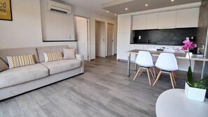Una sala de estar moderna con un sofá, sillas y una mesa de comedor. Hay una zona de cocina con armarios blancos y encimera negra.