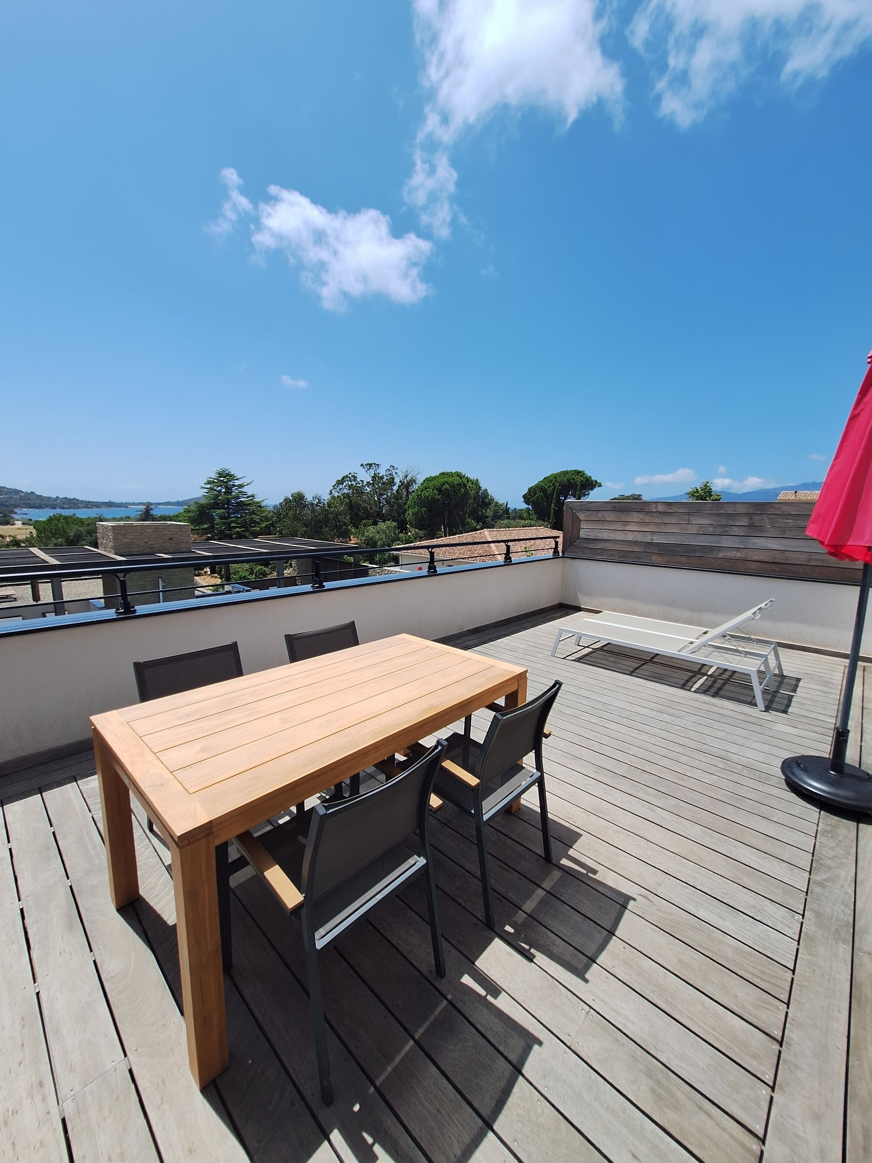 Eine Holzterrasse mit Tisch und Stühlen, einem roten Sonnenschirm und Blick auf die Landschaft.