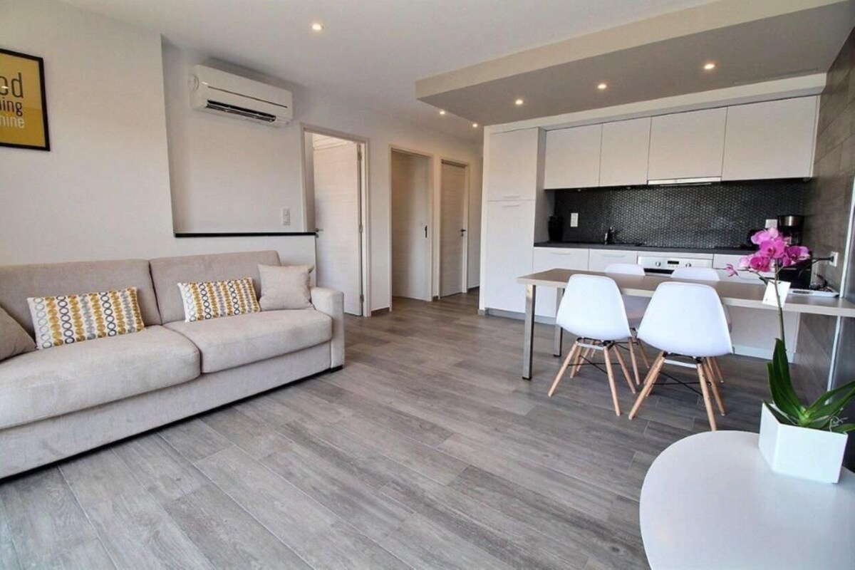Una sala de estar moderna con un sofá, sillas y una mesa de comedor. Hay una zona de cocina con armarios blancos y encimera negra.