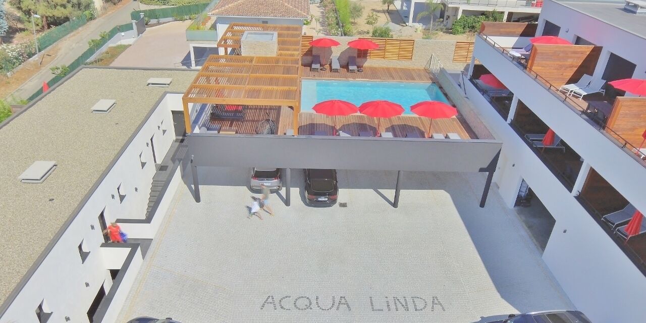 Un espace piscine aménagé sur le toit, avec des parasols et des chaises longues, une enseigne indiquant « ACQUA LINDA », et une vue dégagée sur les bâtiments environnants.