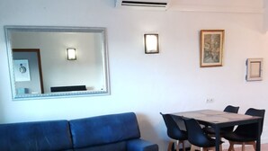 Ein Wohnzimmer mit blauem Sofa, einem weißen Couchtisch, einem Esstisch mit Stühlen und einem großen Spiegel an der Wand.