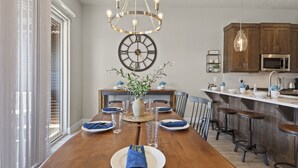 Zona pranzo con tavolo in legno, sedie, un lampadario, un orologio e una cucina sullo sfondo.