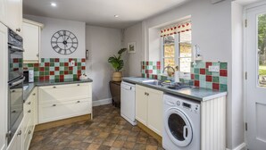 Une cuisine avec des meubles blancs, une grande horloge, un lave-linge et un pare-feu en carreaux.