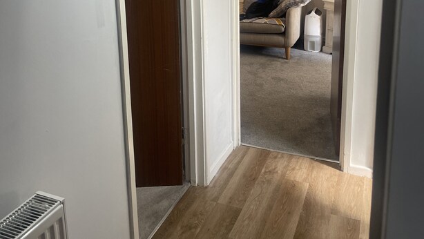 En gang med radiator, trægulv og en dør, der fører til et opholdsområde med en sofa og et lille bord.