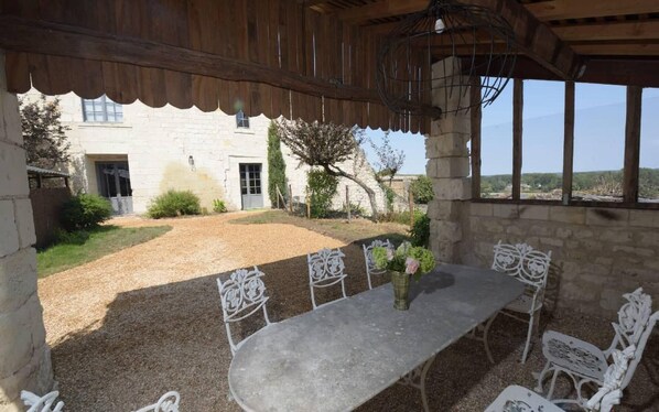 A covered outdoor dining area with a table and chairs, a stone building in the background, and a view through the window. - When heritage, art and well-being meet (CANDES SAINT MARTIN)