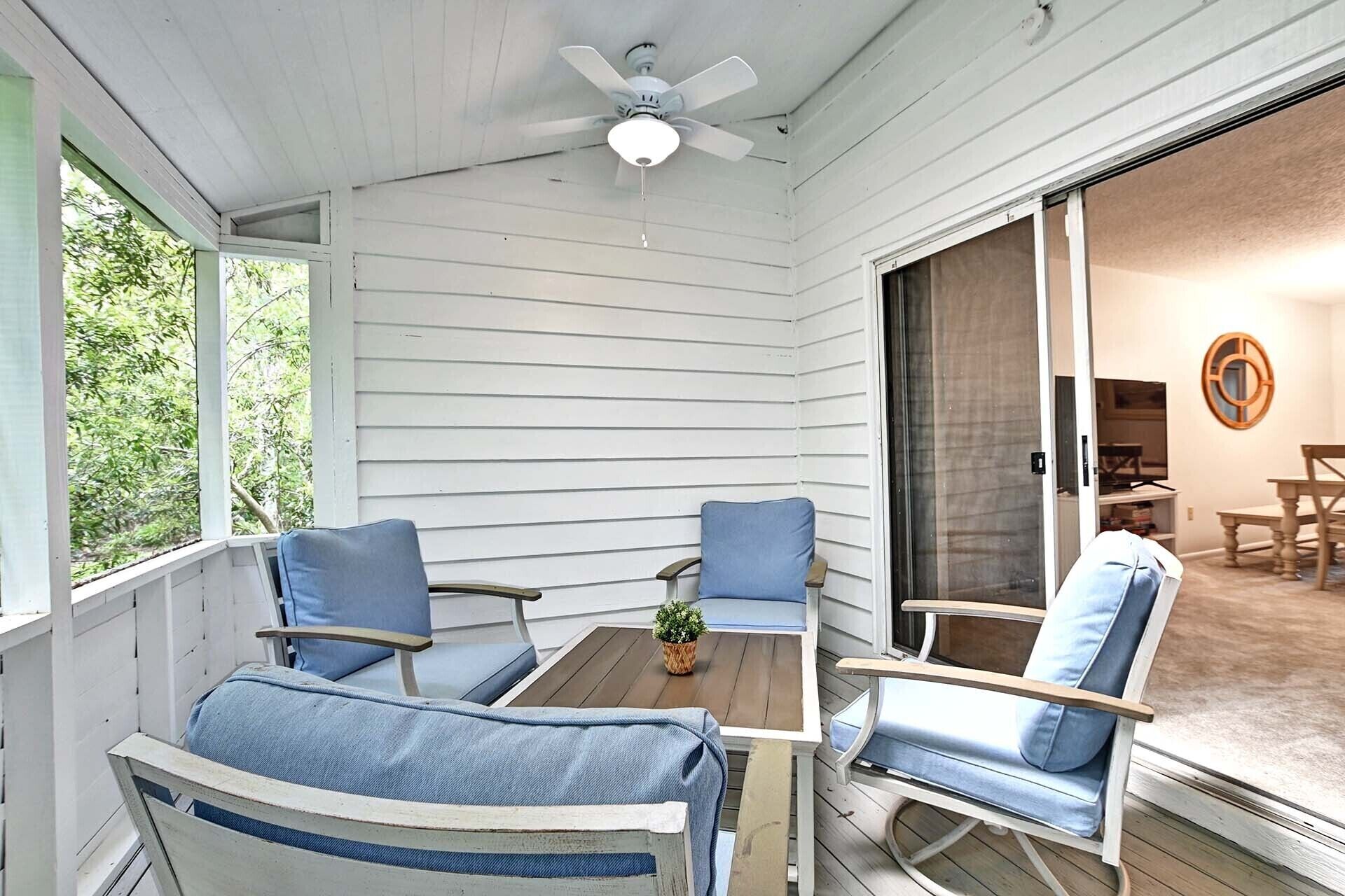 A covered porch with two blue cushioned chairs, a wooden table, and a ceiling fan.