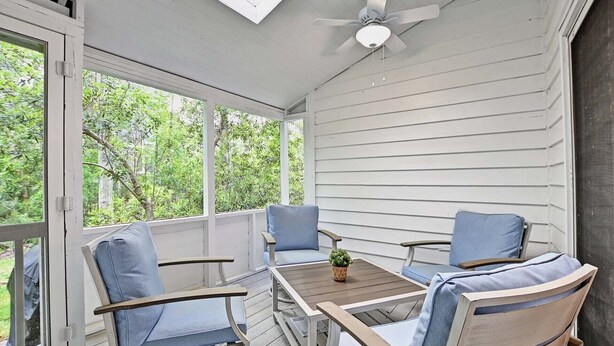 A covered porch with two chairs and a table, large windows, and a ceiling fan.
