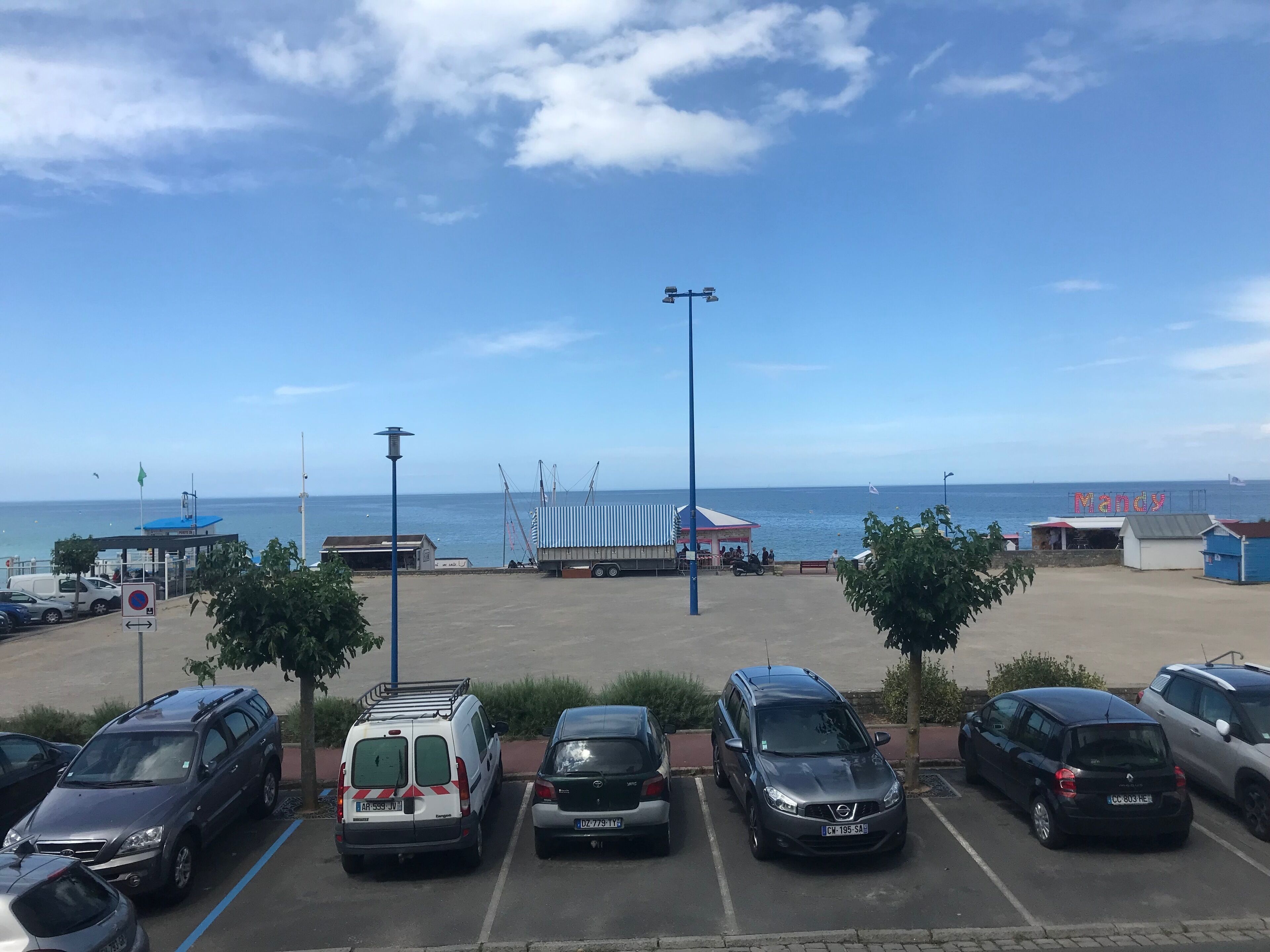 A parking lot with several cars, a small building with a blue roof, and a view of the sea.
