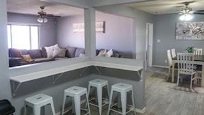 En moderne stue med en grå sofa, en hvid bardisk med barstole og et spiseområde med et træbord og stole.