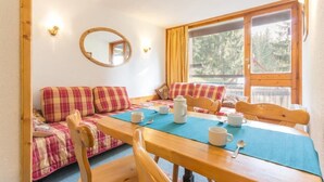 Una habitación acogedora con una mesa de madera, sillas, un sofá con cojines de cuadros, un espejo y una ventana con cortinas.
