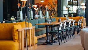 Een moderne eetruimte met houten tafels en stoelen, een bank met gele kussens en hangende hanglampen.