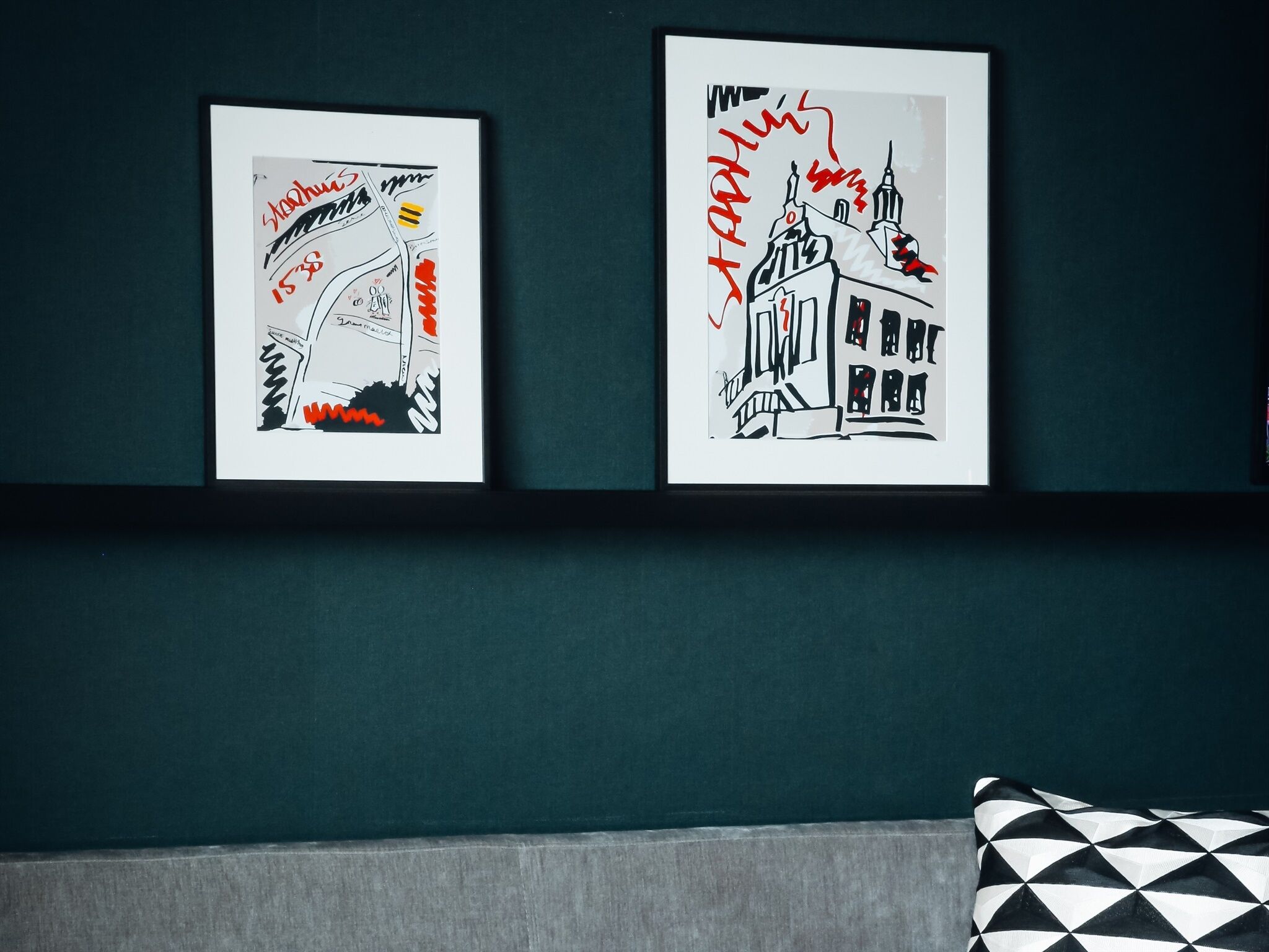 Two framed artworks on a dark teal wall, one depicting a building with red text and the other a building with black and white text.