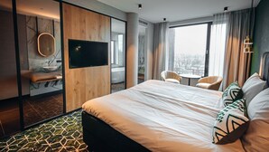 Ett modernt hotellrum med en stor säng, en TV, en sittgrupp med stolar och utsikt över staden genom stora fönster.