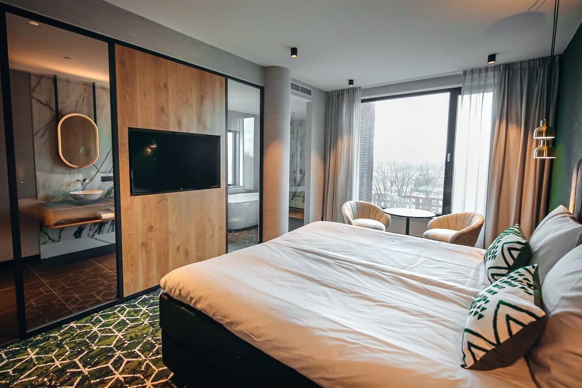 Une chambre d’hôtel moderne dotée d’un grand lit, d’une télévision, d’un coin salon avec des fauteuils et offrant une vue sur la ville grâce à de grandes fenêtres.