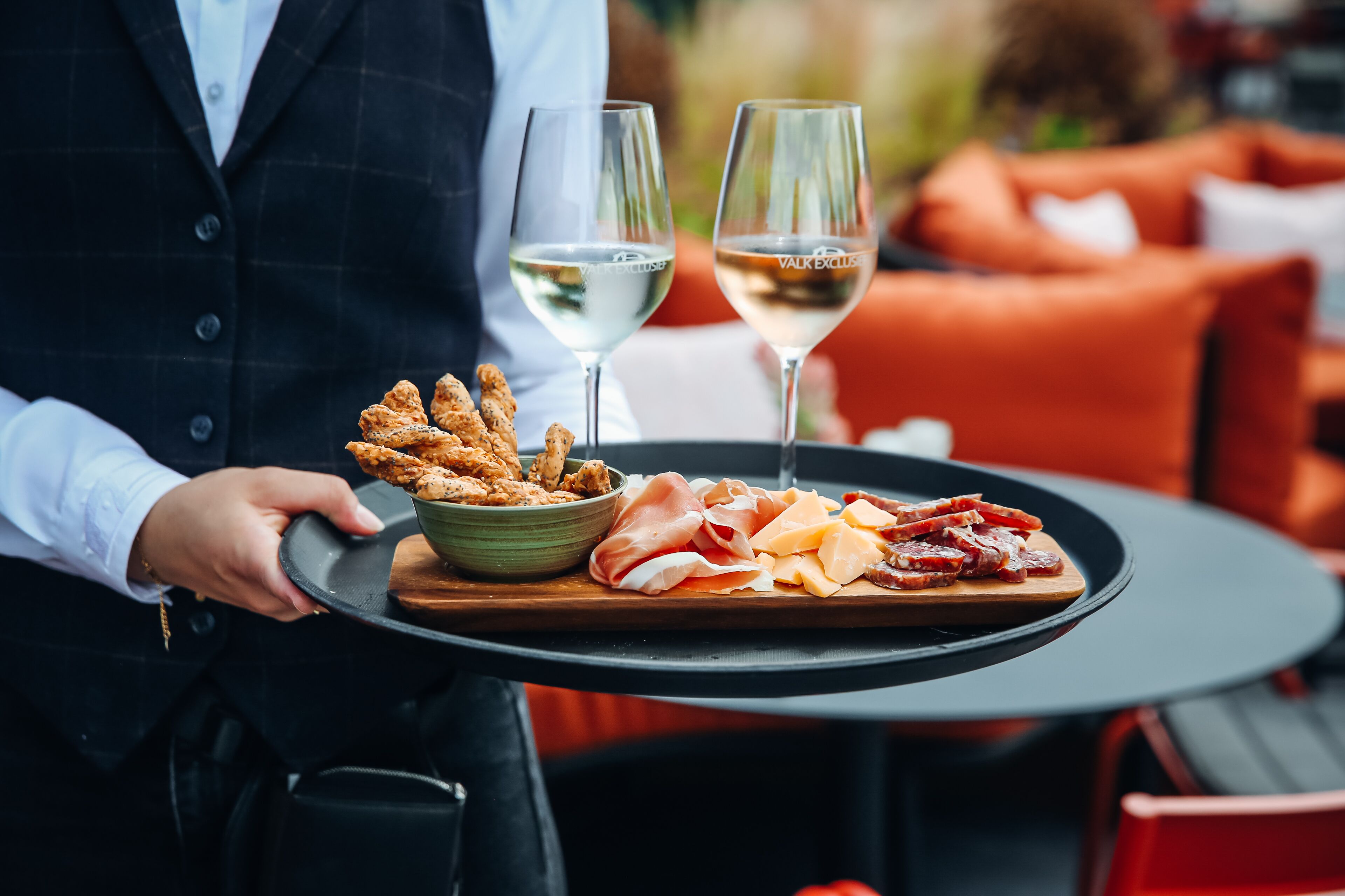 Un camarero sostiene una bandeja con palitos de pan, un plato de embutidos y dos copas de vino blanco.