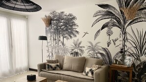 Una sala de estar con un sofá color beige, una mesa de centro de madera, una mesita auxiliar y un mural de follaje tropical.