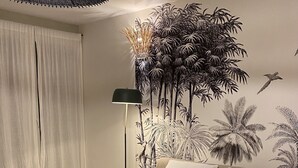 Un dormitorio con una cama grande, dos almohadas, una lámpara de pie y un mural en la pared con palmeras y un pájaro.